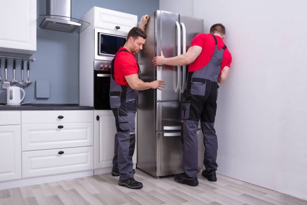 two young male movers in uniform placing modern steel refrigerator in kitchen